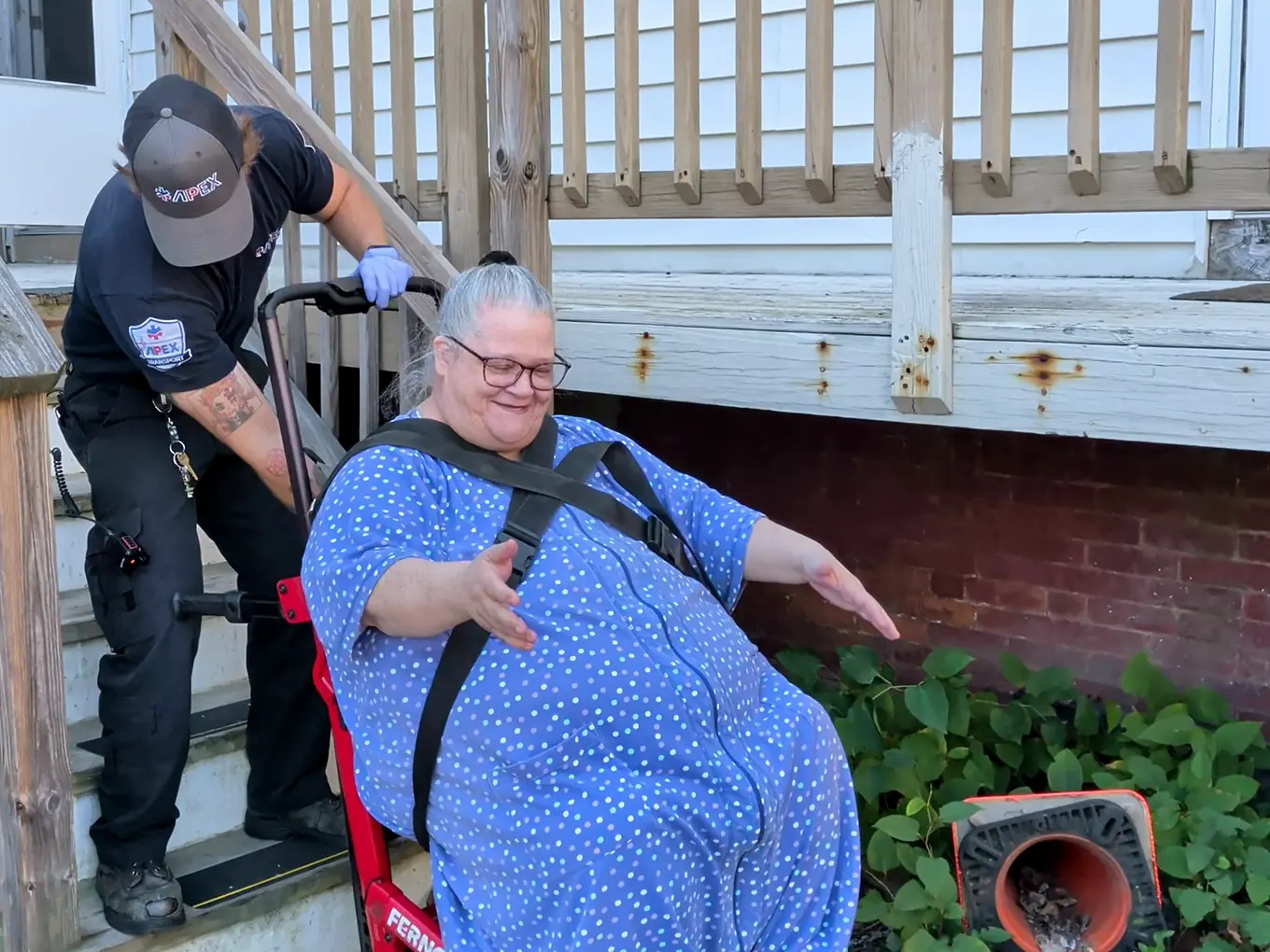 A smiling rider reaches the bottom of her porch steps in a red stair chair guided by an Apex crew member